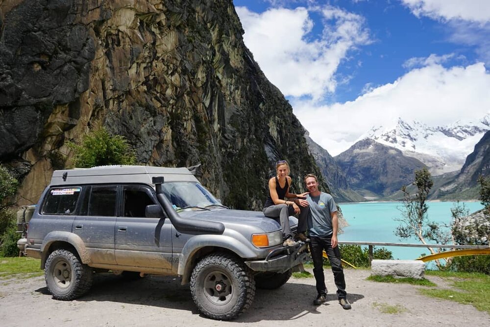 Man and woman leaning on their overland vehicle in from of a glacial lake.