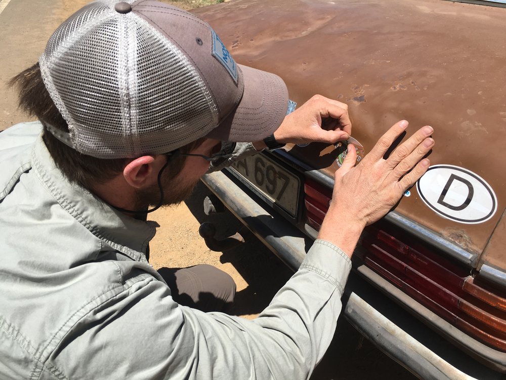 Isaac applies an Overland Expo decal at Blue's request. This will be the final addition before the Benz is entombed in a museum.