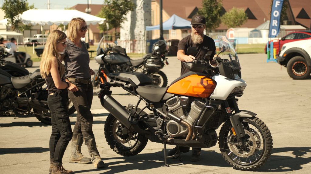 Two women motorcycle riders learn about the Harley Davidson Pan America from a man with a beard.