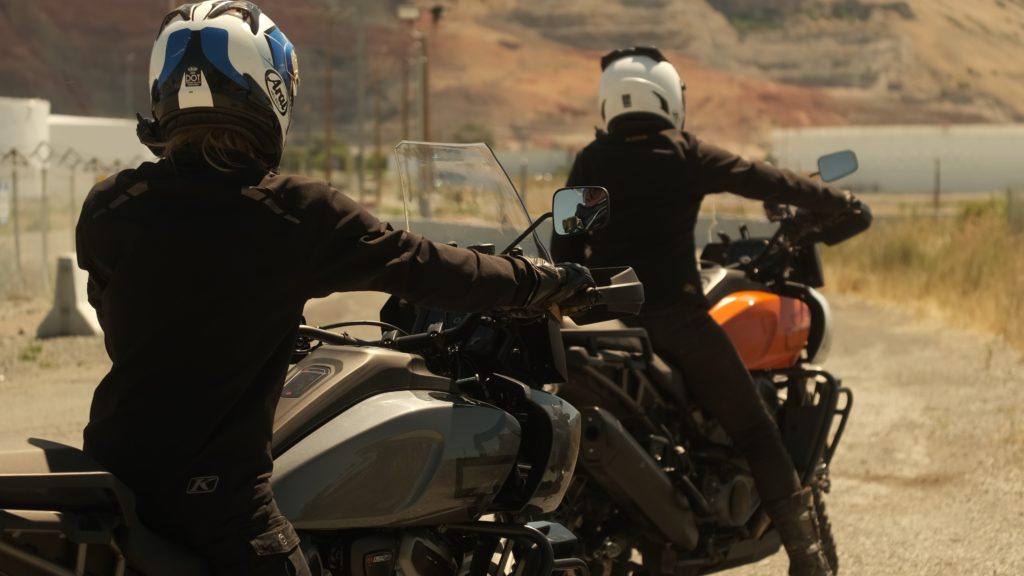 Two women ride away on motorcycles on a gravel road