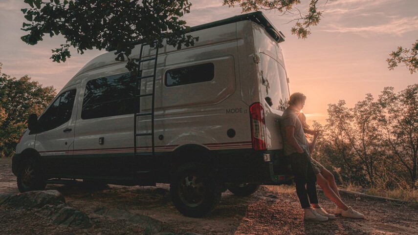 Couple leaning against the back of a Storyteller Overland MODE LT at a dispersed campsite in the woods.