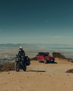 A woman rides a Honda Africa Twin motorcycle away from campsite followed by a Toyota 4Runner