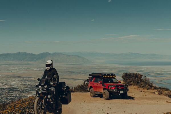 A woman rides a Honda Africa Twin motorcycle away from campsite followed by a Toyota 4Runner