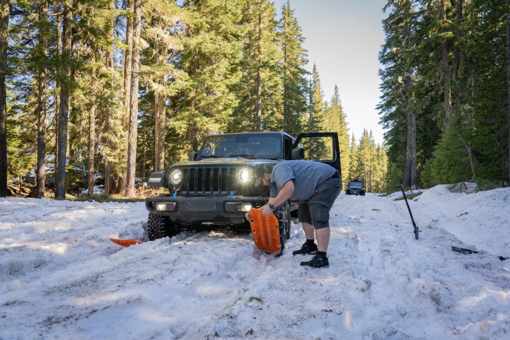 A man is digging out snow beneath the tires of a Wrangler with MAXTRAX