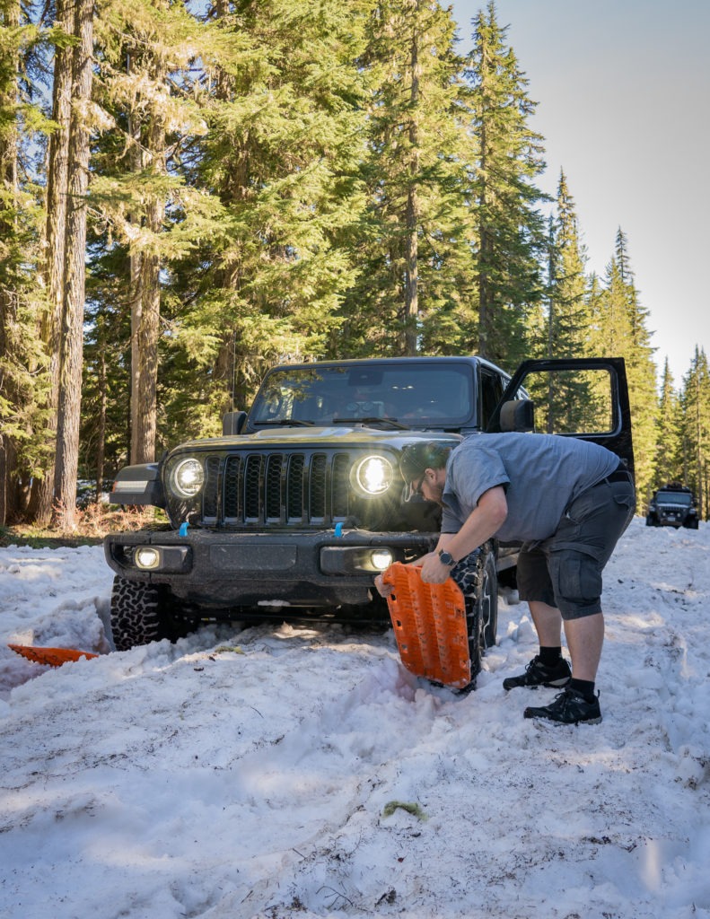 A man is digging out snow beneath the tires of a Wrangler with MAXTRAX