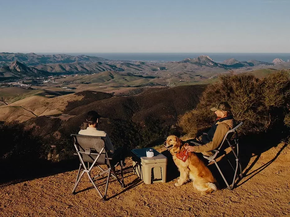 Two people and a dog taking in the view from camp in a YETI TrailHead Camp Chair. 