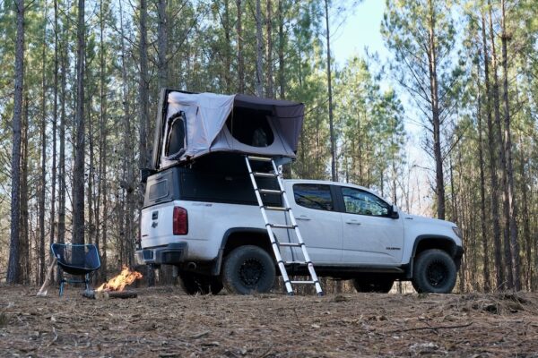 Campsite amongst pine trees with a rooftop tent on a truck, a camp chair, and campfire.