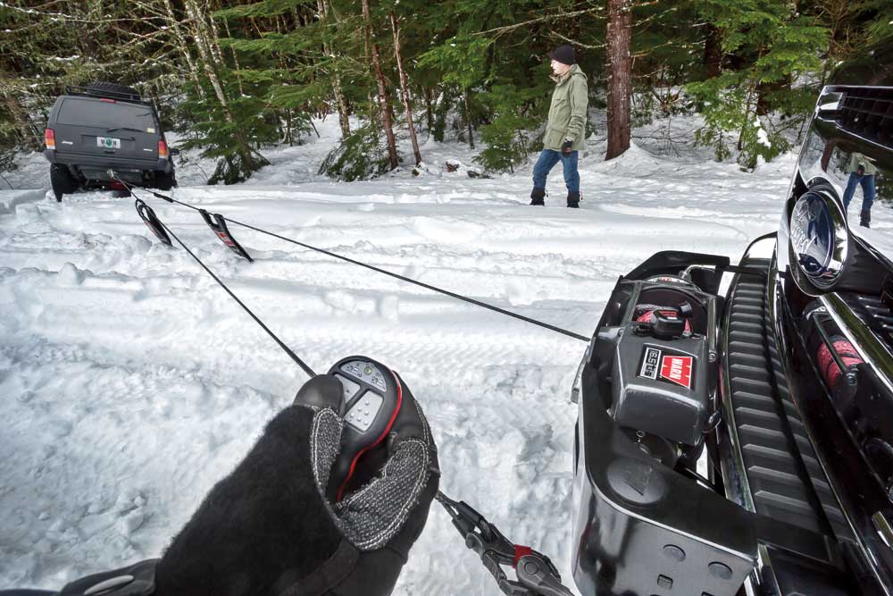 People using a winch to extract a vehicle from a ditch in a snowy forest.